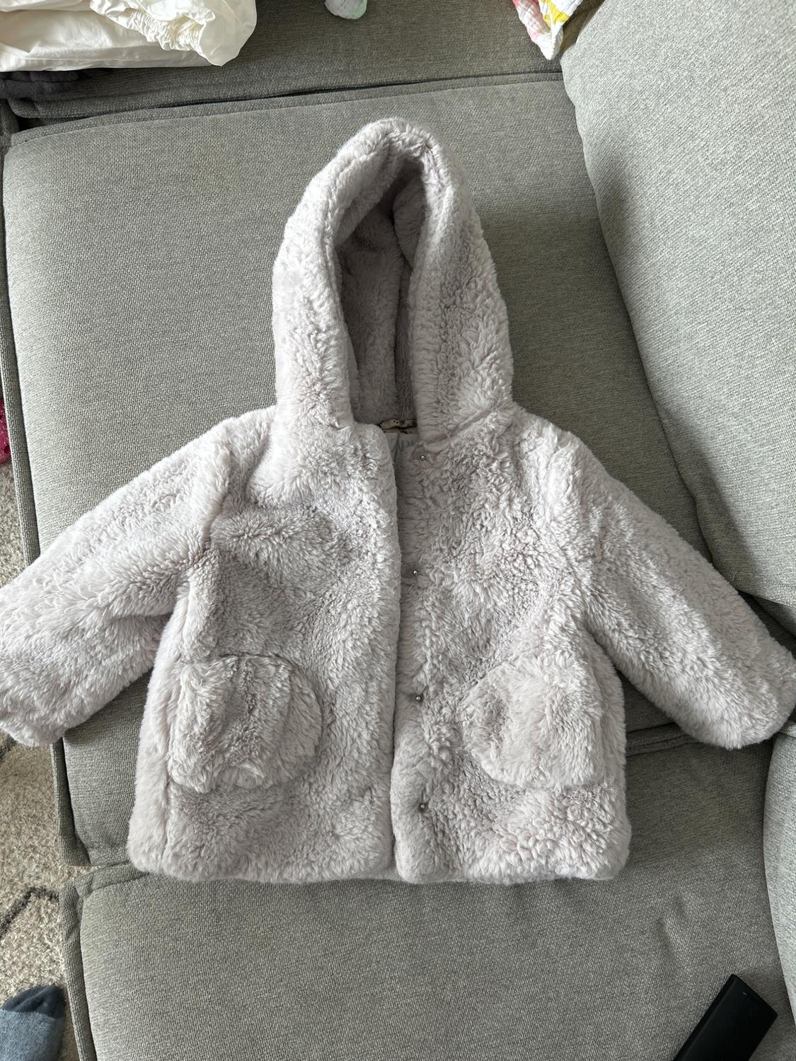Light grey plush jacket with a hood and round pockets, placed on a grey sofa.