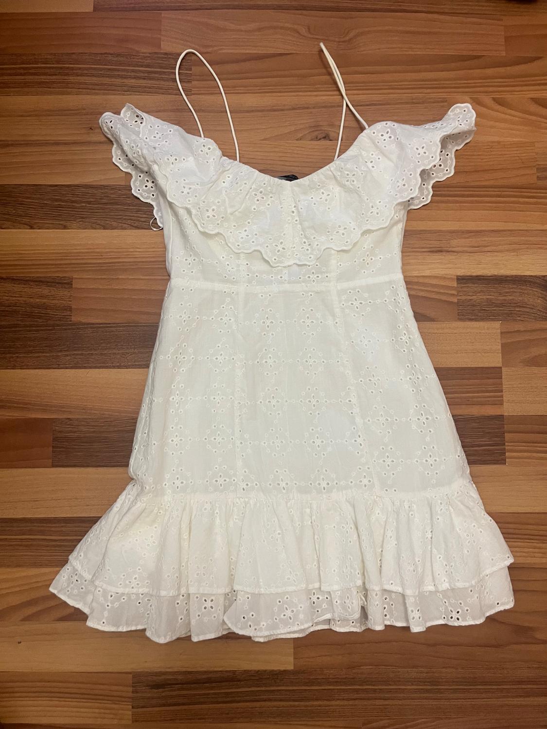 White spaghetti strap dress with ruffles and openwork embroidery, on a brown wooden background.