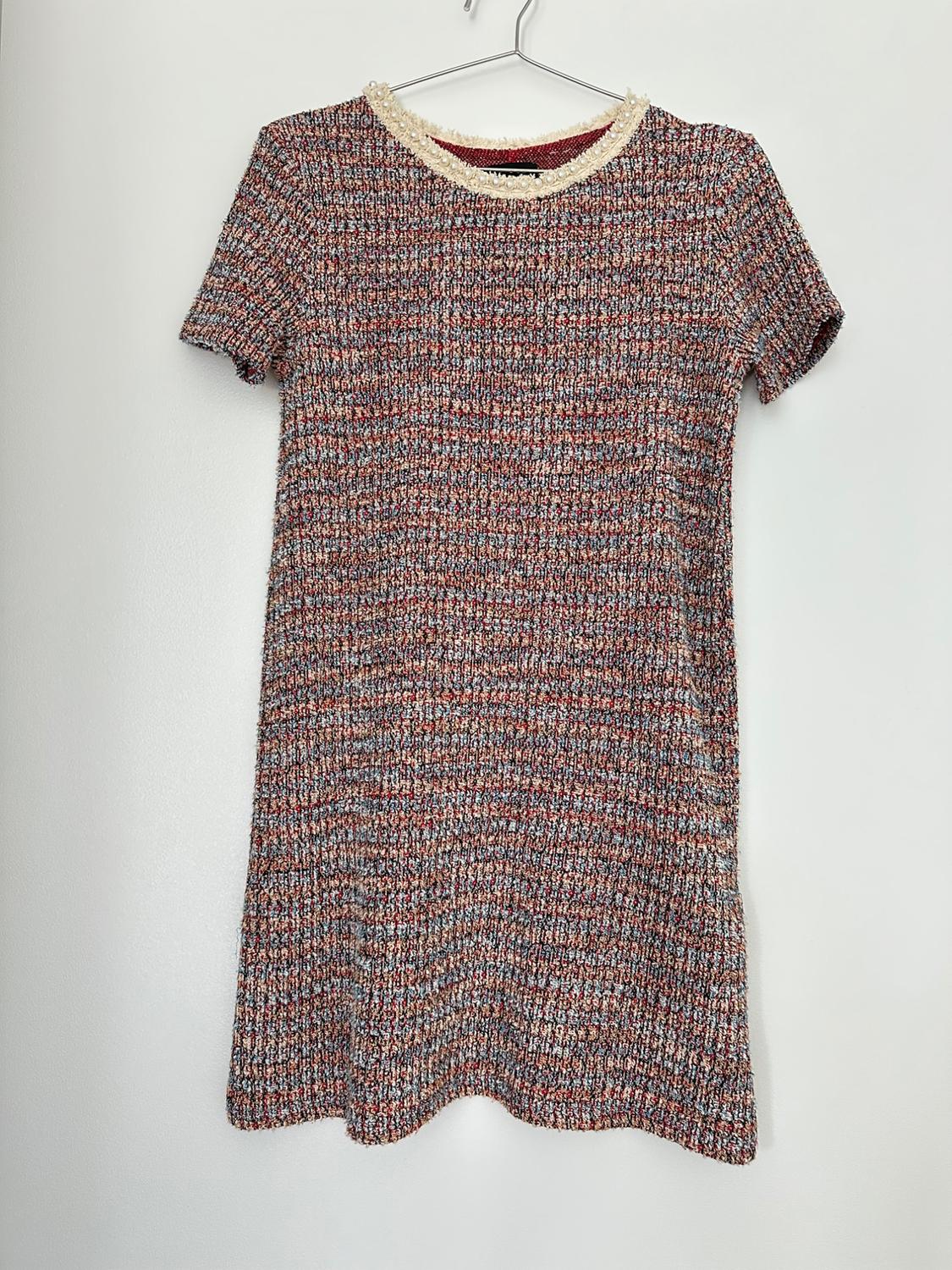 Multicolor textured short dress, short sleeves, round neckline with pearls and frayed hem, hanging on a hanger against a white wall.