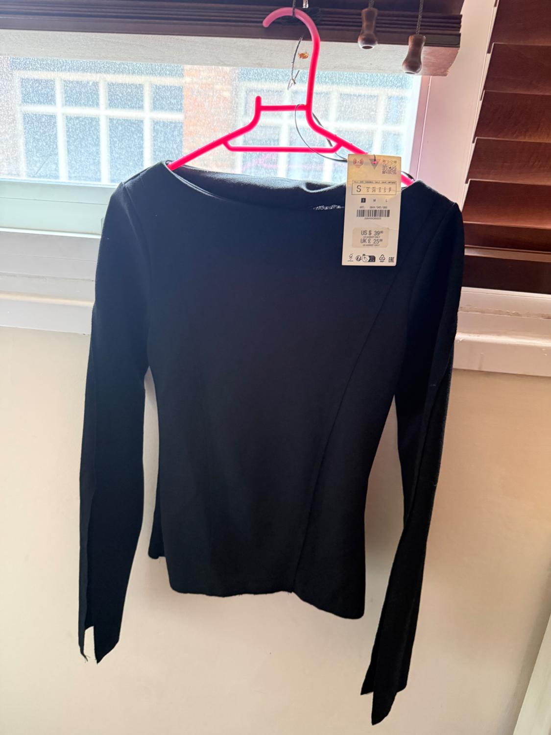 Black long-sleeved top hanging on a pink hanger in front of a window. Label with text: S US $ 39 UK £ 25.
