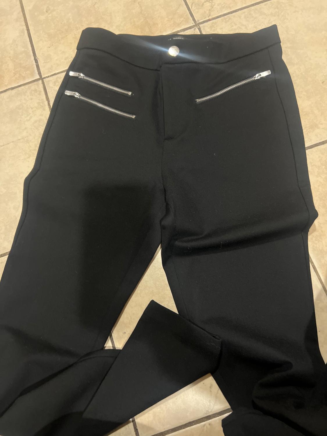 Black trousers with silver zips on the front pockets, laid on a beige tiled floor.