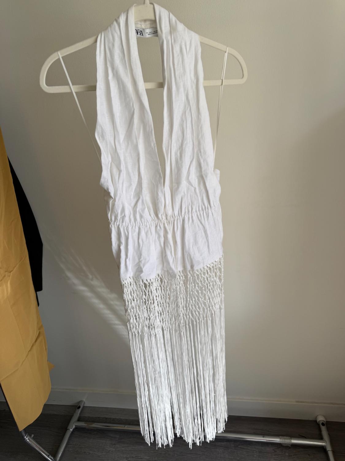 White dress hanging on a hanger, with a deep neckline and long fringes at the bottom. White wall background.