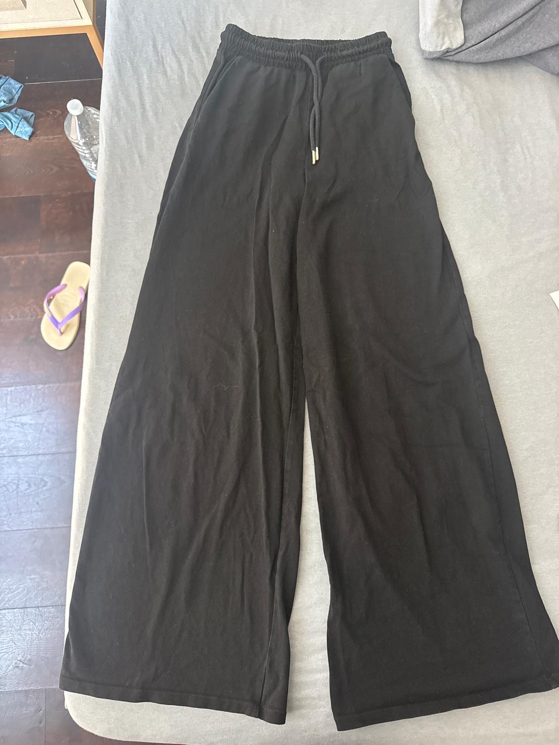 Black wide-leg trousers with an elasticated drawstring waist, placed on a grey bed. In the background, a bottle and a sandal.