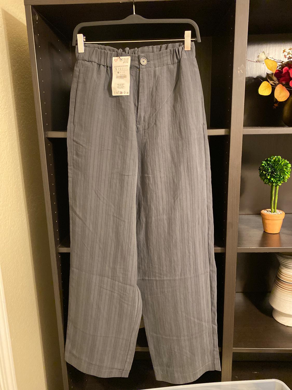 Light grey fabric trousers with a swing tag, hanging on a black hanger on a dark shelf with plant decoration.
