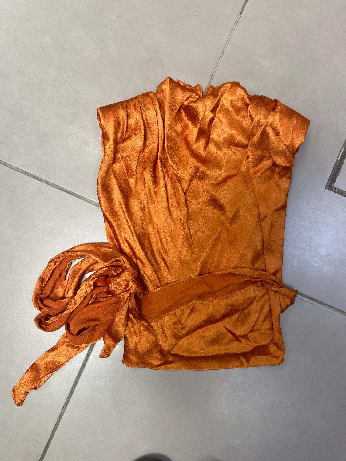 Sleeveless satin orange top with a tie detail at the waist, placed on a grey tiled floor.