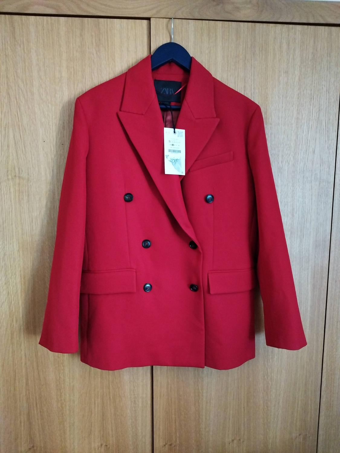 Red double-breasted jacket hanging on a black hanger in front of a light wooden wardrobe. Label visible on the collar.