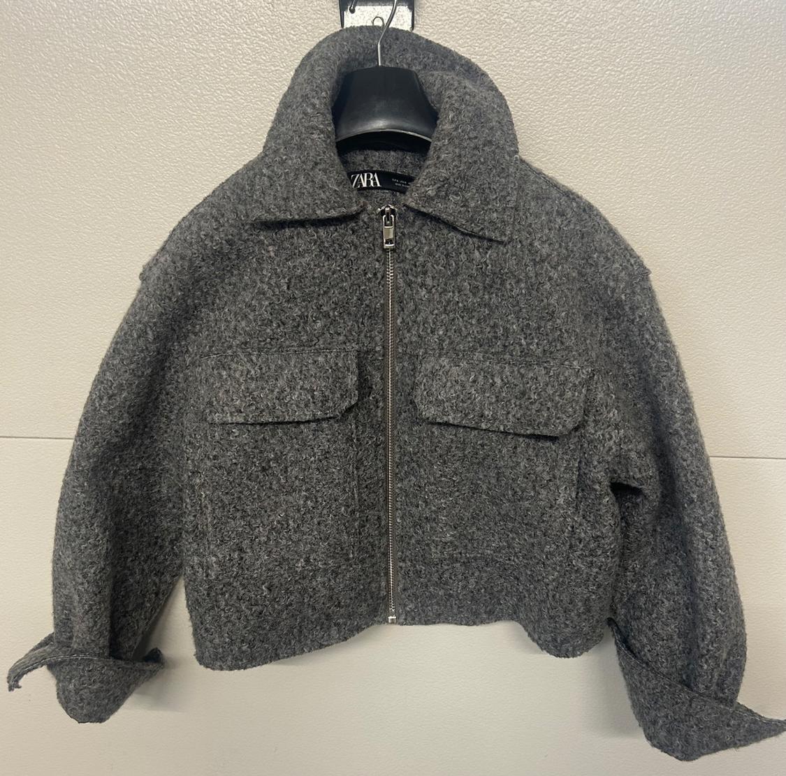 Textured grey cropped jacket with zip fastening and flap pockets, hanging on a hanger against a white wall. ZARA label.