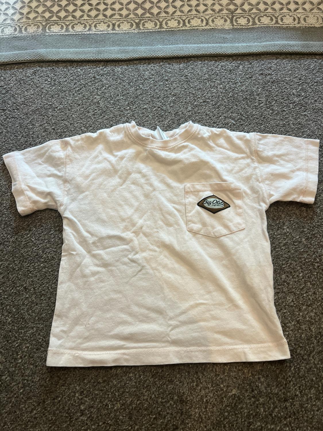 Short-sleeved T-shirt in a light shade on a grey rug. It features a chest pocket with a patch that says "Big Olita".