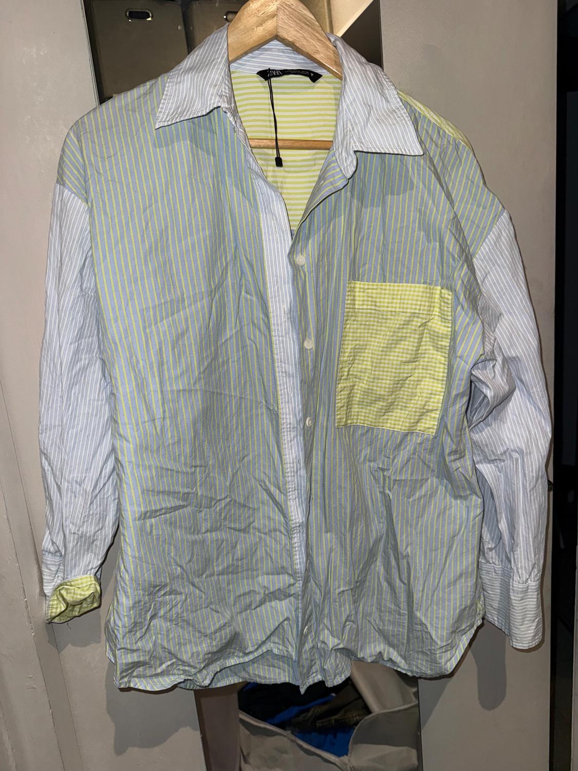 Vertical striped shirt in shades of blue, green, and yellow, hanging on a wooden hanger against a neutral background.