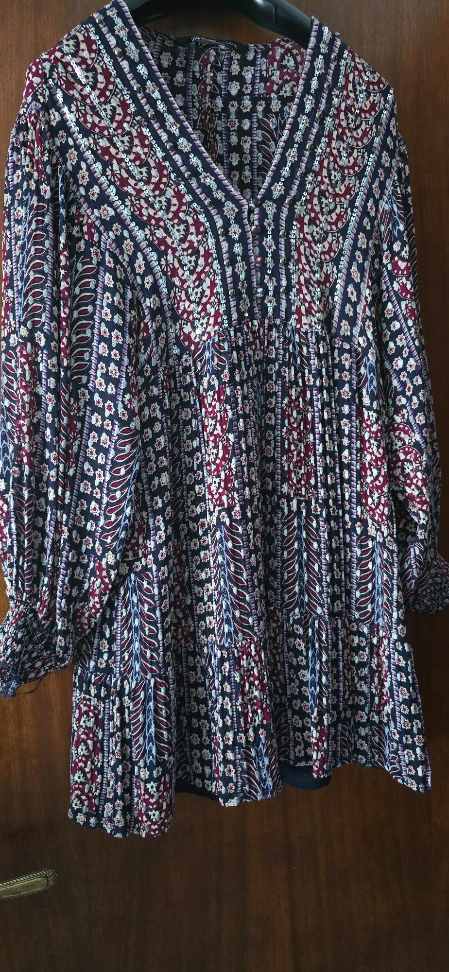 Long-sleeved blouse with a floral print in shades of blue, white, and burgundy, hanging on a hanger against a wooden background.