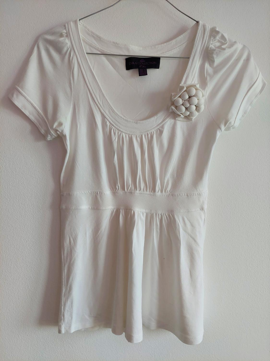 White short-sleeved T-shirt on a hanger against a white wall. Round neck, gathers, floral appliqué, and label with ZARA COLLECTION text.