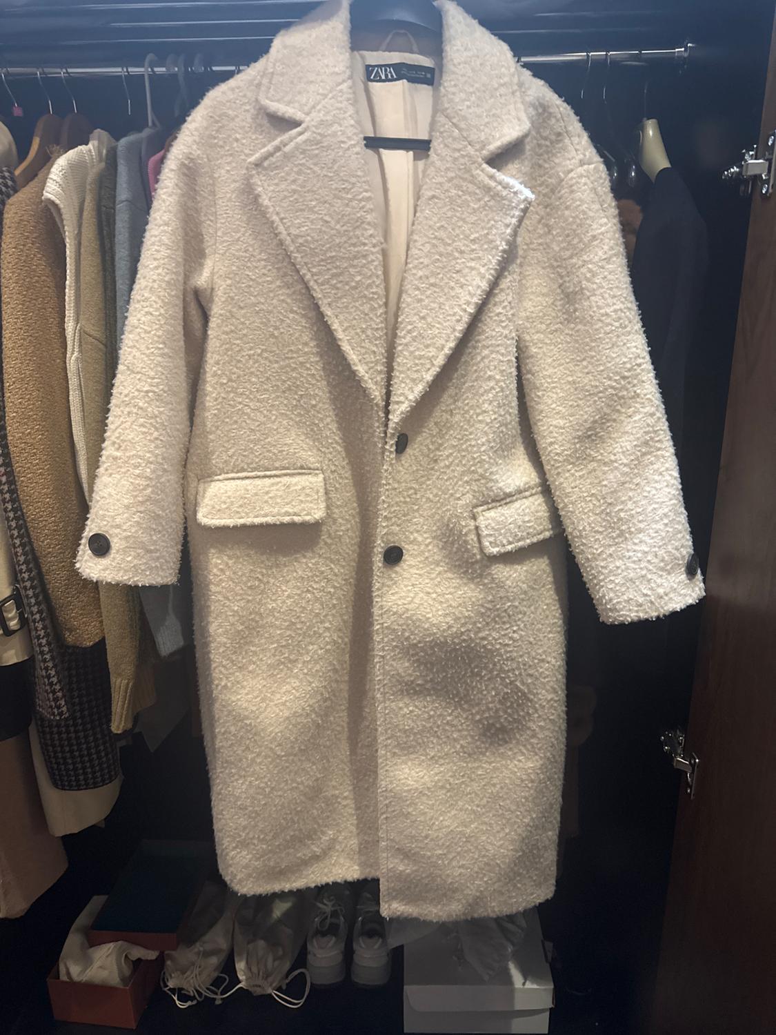 White wool coat with lapels and black buttons hanging in a wardrobe, surrounded by other garments and boxes on the floor.