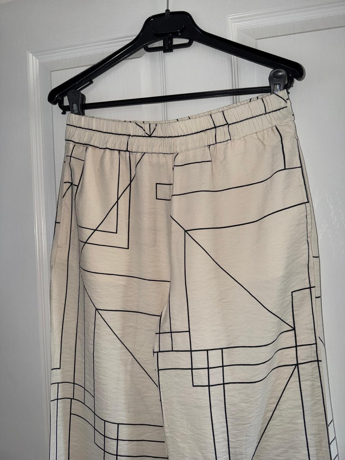 Beige trousers with a black geometric print, hanging on a black hanger over a white door.