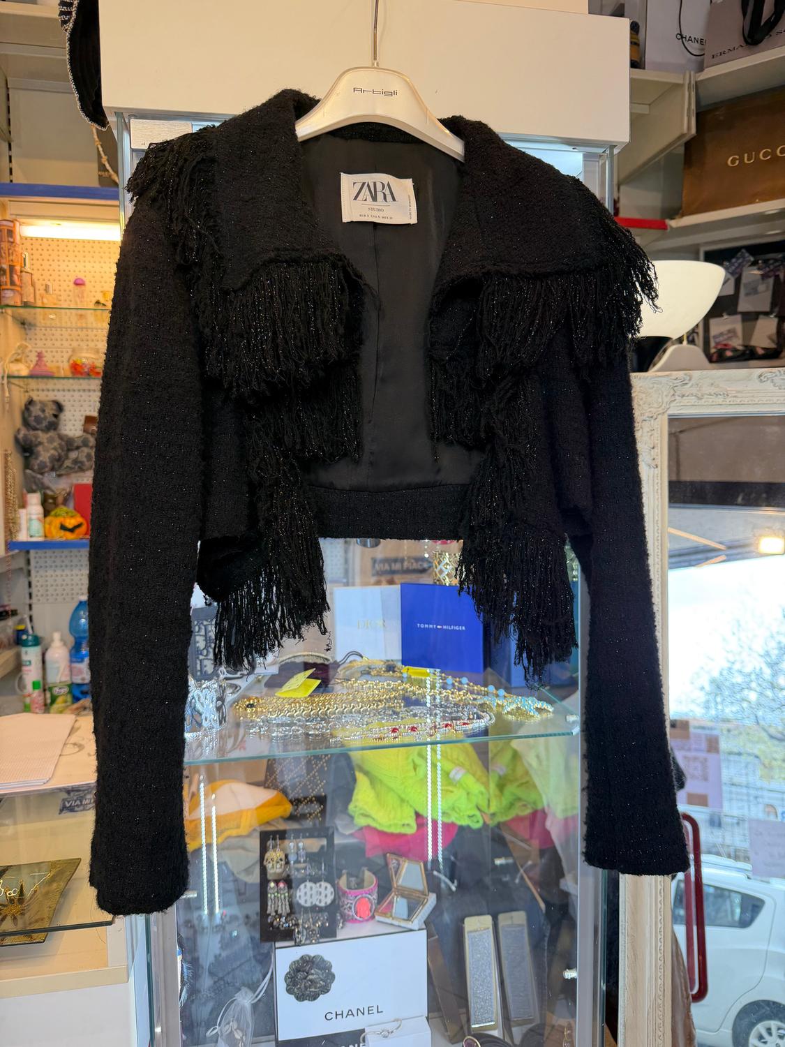 Black fringed jacket hanging on a white Zara Studio hanger, displayed in a glass cabinet with accessories and clothing in the background.