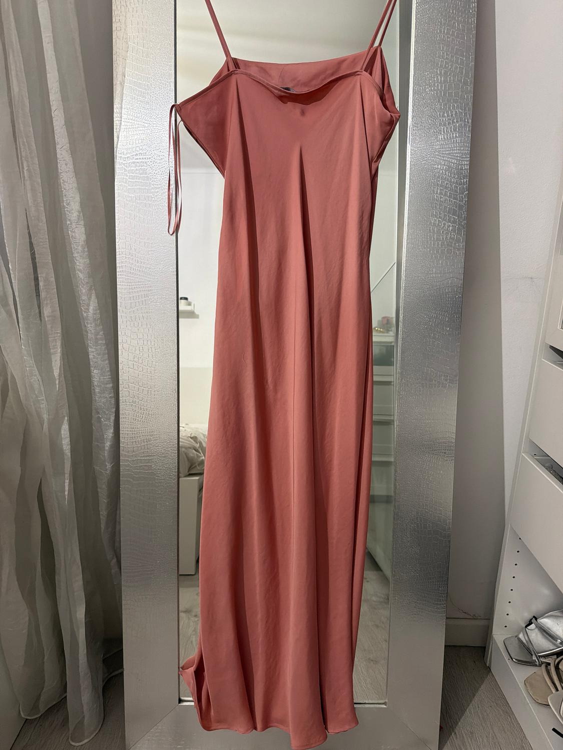 Long pink dress hanging on a full-length mirror, with thin straps and a background of a white room with curtains.