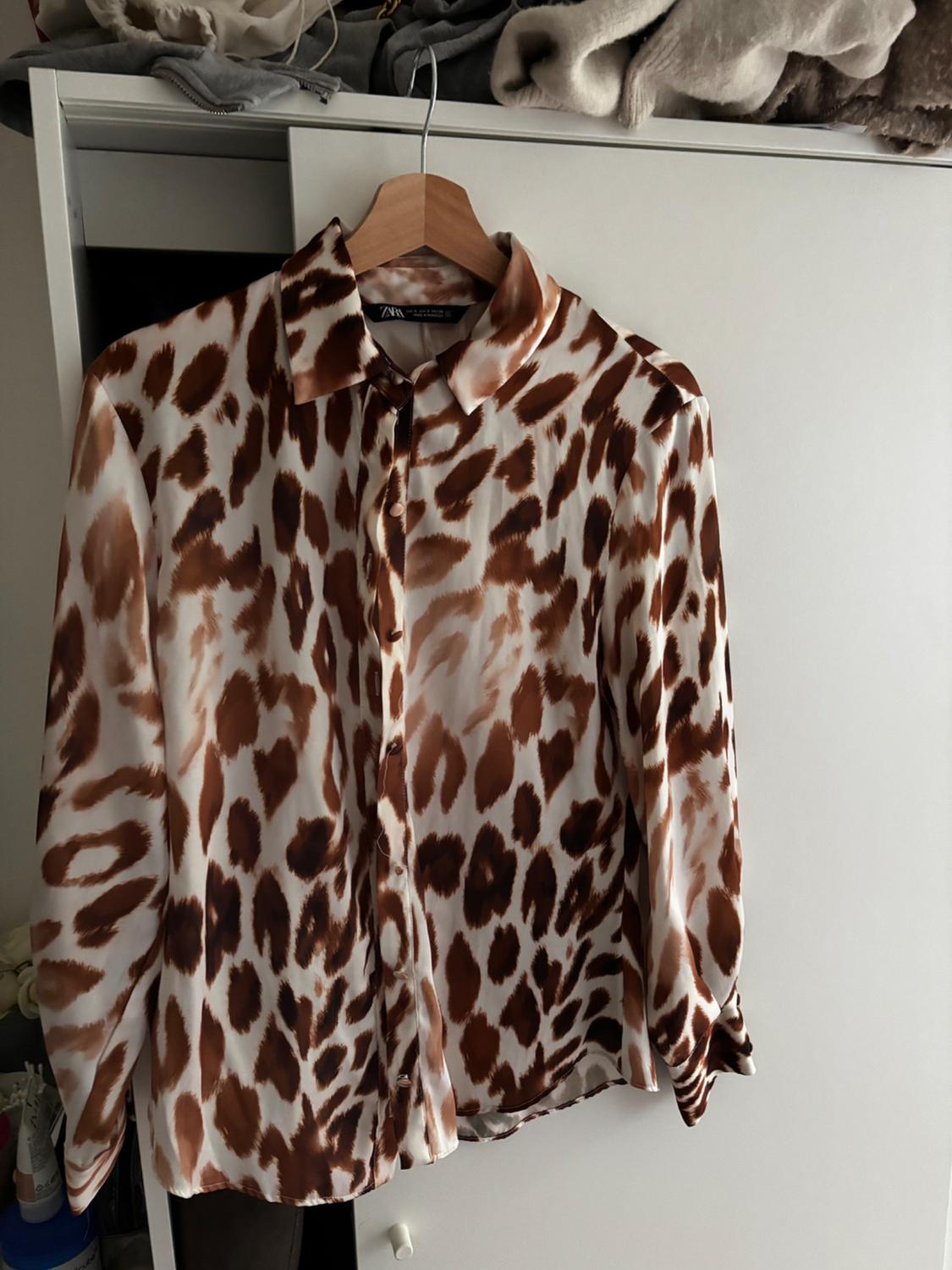 Brown and white animal print long-sleeved shirt hanging on a wooden hanger in a white wardrobe. Visible text: ZARA.
