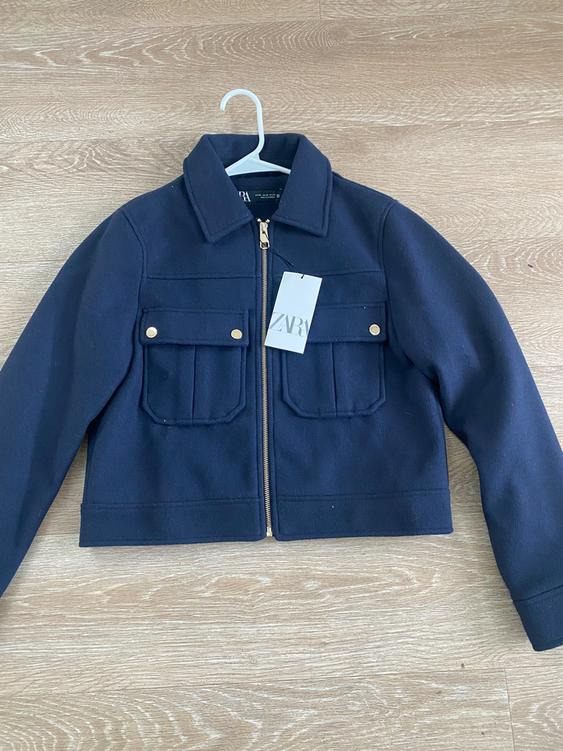 Navy blue jacket with a gold zipper and front pockets, hanging on a white hanger over a light wood floor. "ZARA" tag.