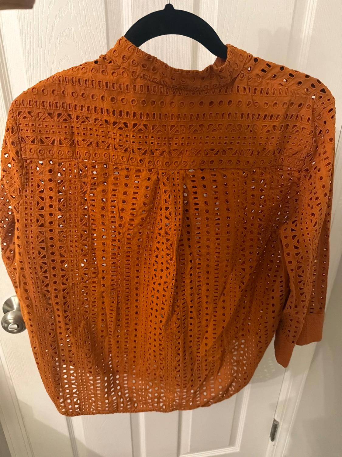 Orange lace blouse with an openwork design, hanging on a black hanger in front of a white door.