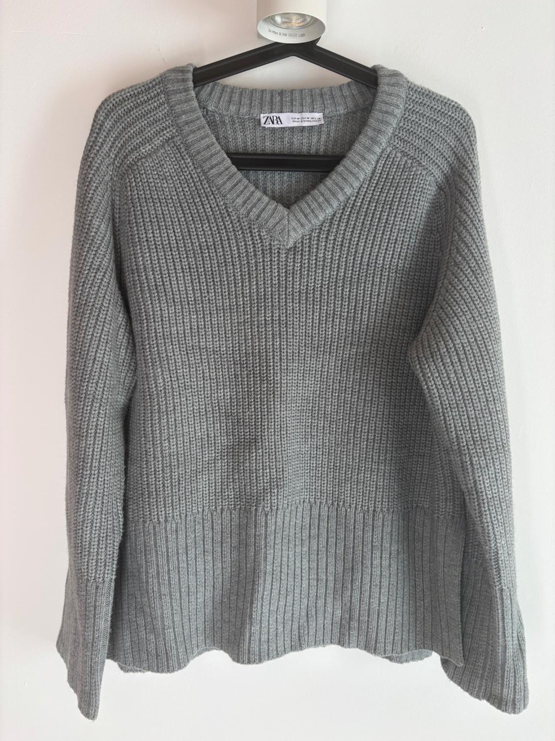 Grey chunky knit V-neck jumper, hanging on a black hanger. Visible label with text "ZARA". White background.