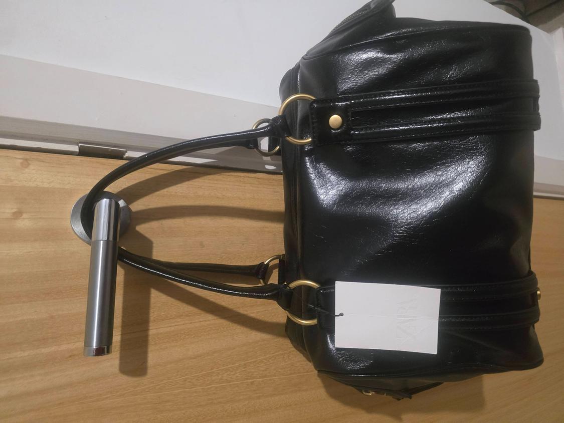 Shiny black handbag hanging from a silver doorknob on a wooden door. It has gold details and a white tag with the text ZARA.