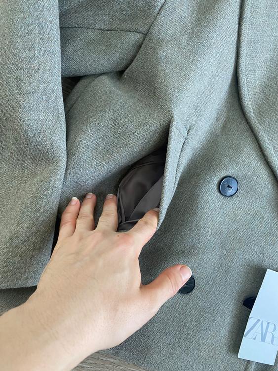 Hand checking the pocket of a gray coat with black buttons. Visible label with the text "ZARA" in the bottom right corner.