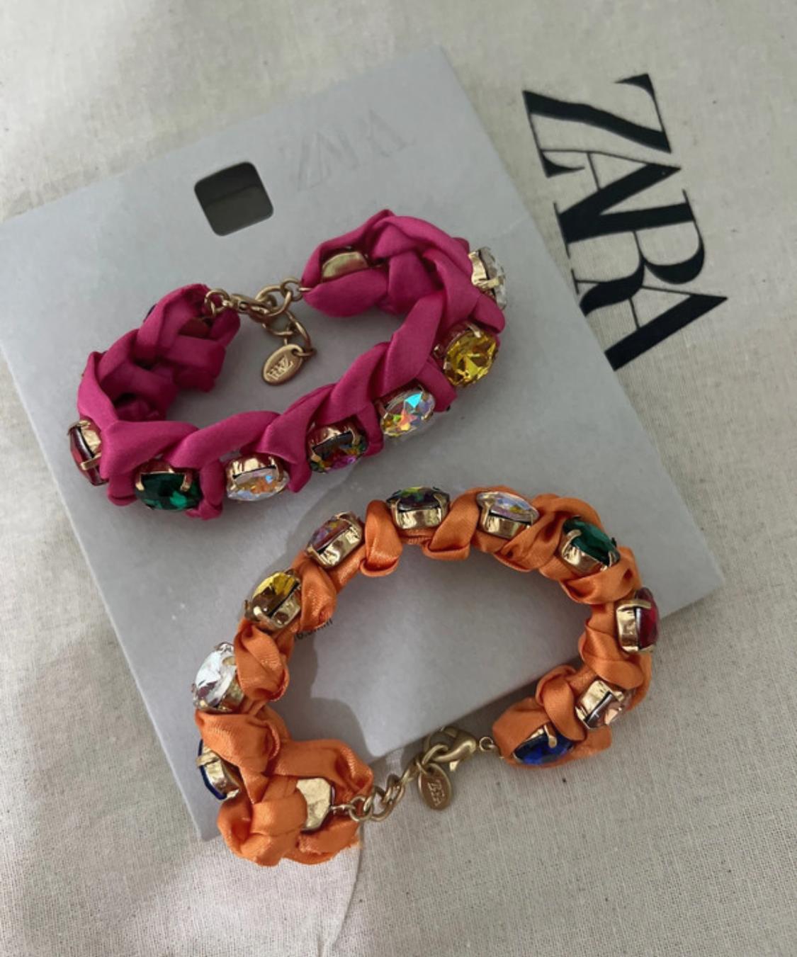 Two braided bracelets with colourful beads, one pink and one orange, on a grey tag and a fabric background with the text ZARA.