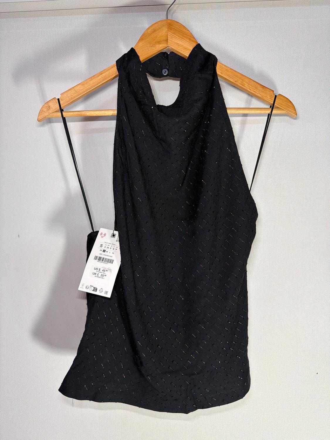 Black sleeveless textured top, hanging on a wooden hanger. Visible tag with sizes and prices in dollars and pounds.