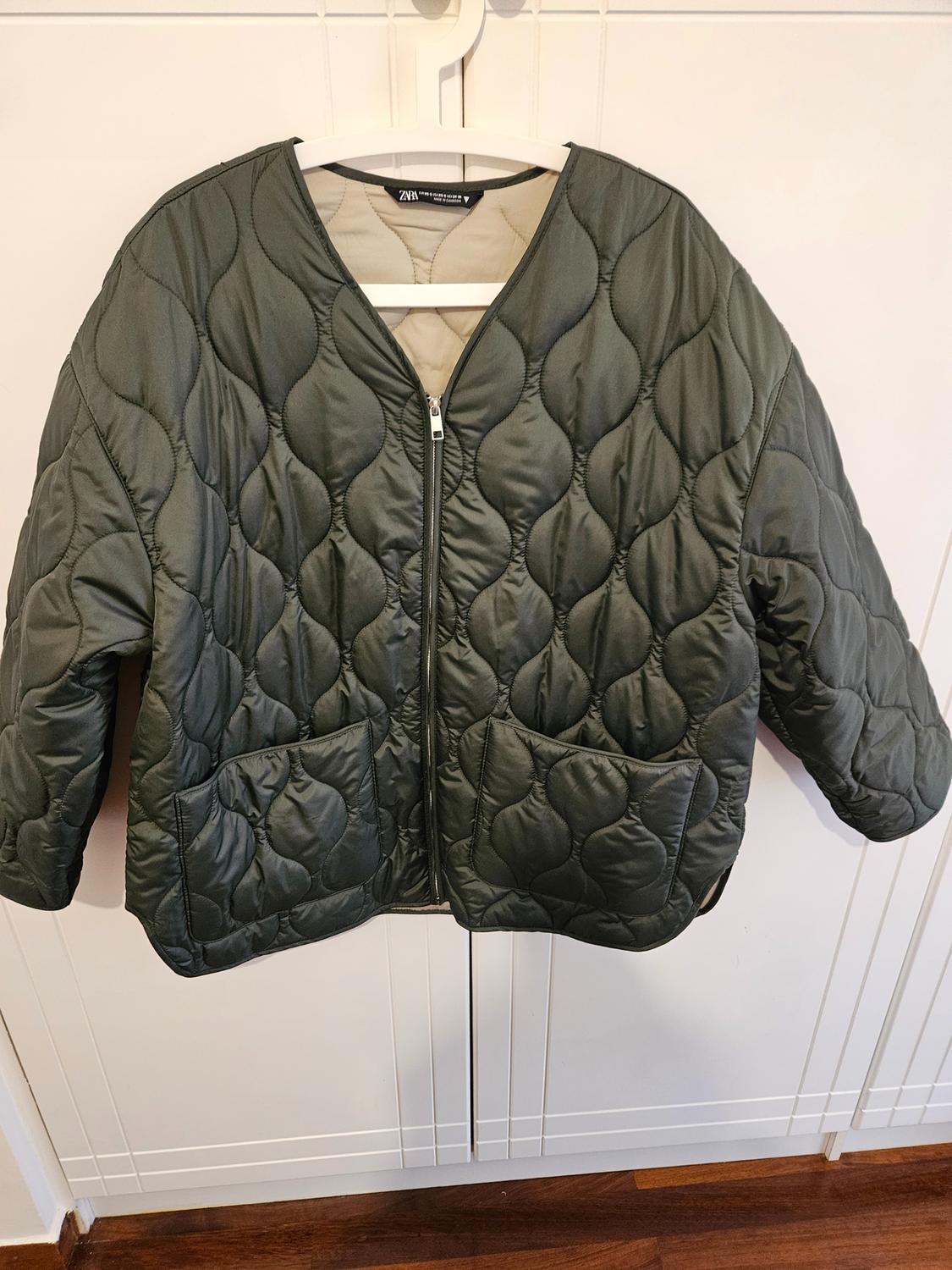 Olive green padded jacket with zip and front pockets, hanging on a white hanger against a white background.