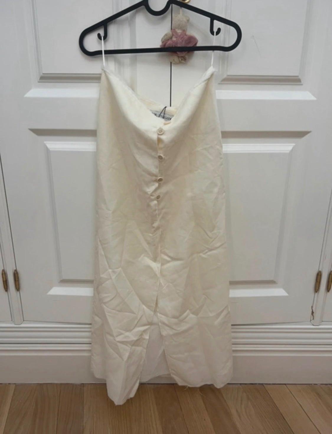 White skirt with front buttons and a slit, hanging on a black hanger over a white door. Relaxed atmosphere with wooden flooring.