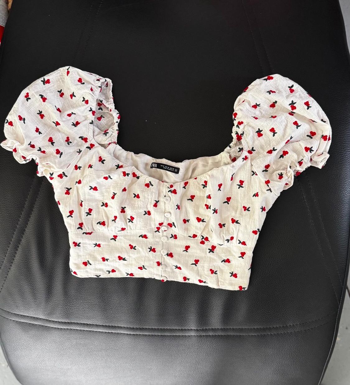 White cropped top with red print, puff sleeves and front buttons, on a black seat. ZARA text on the label.