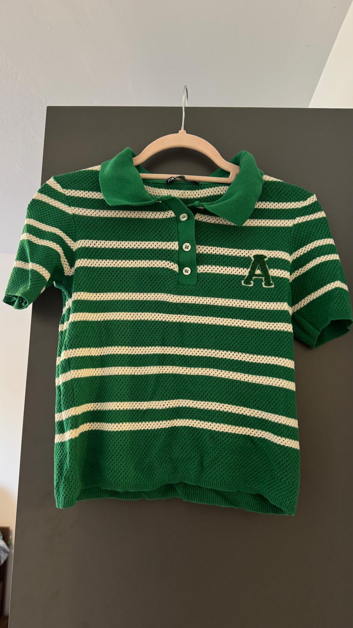 Green polo shirt with white stripes and an embroidered "A" on the chest, hanging on a wooden hanger against a grey background.