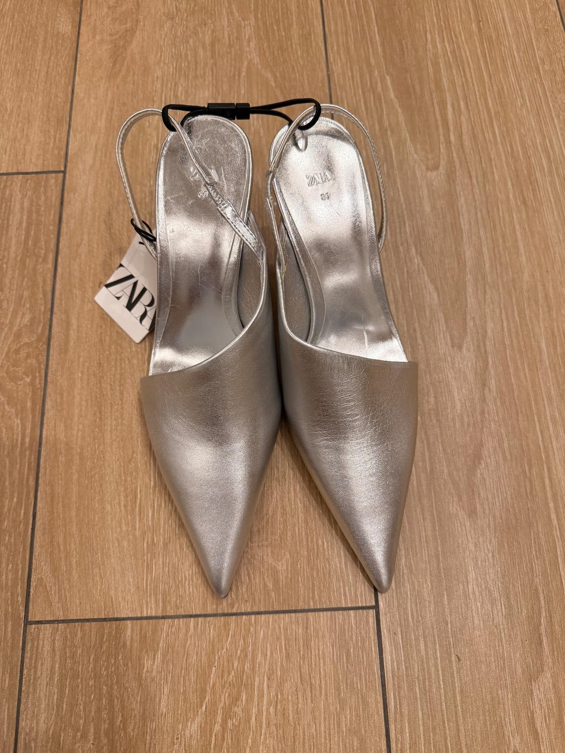 Silver pointed shoes with thin straps, on a light wooden floor. Visible label with the text "ZARA".