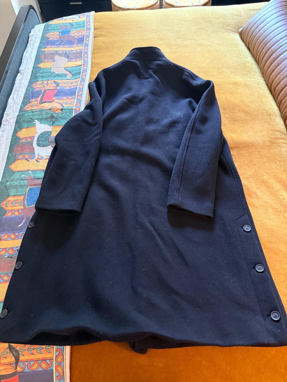 Navy blue long coat with buttons on the side hem, on a bed with a mustard bedspread and patterned fabric. Relaxed composition.