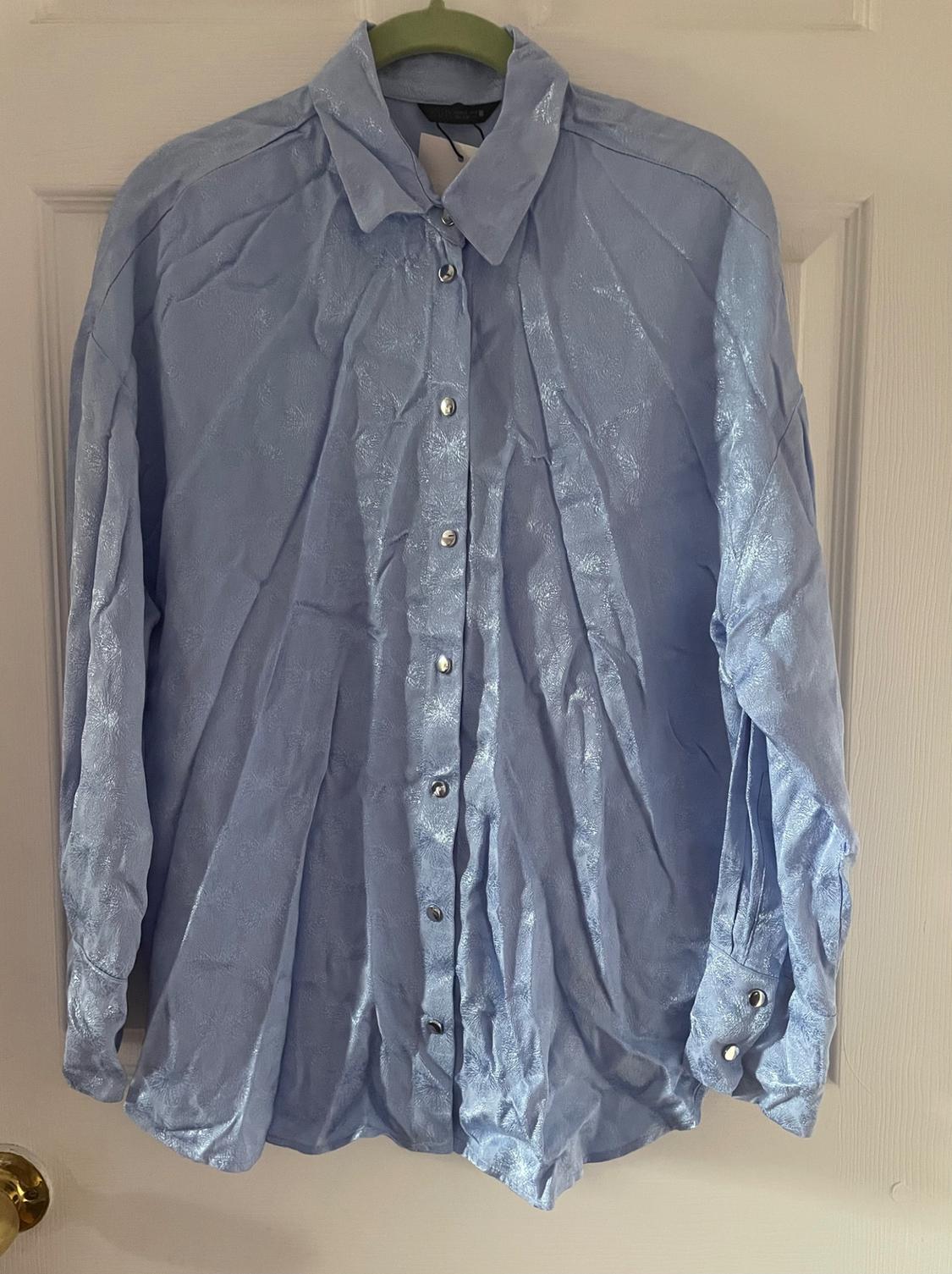 Light blue shirt with a shiny texture, hanging on a green hanger. It has silver buttons and is creased. White door background.