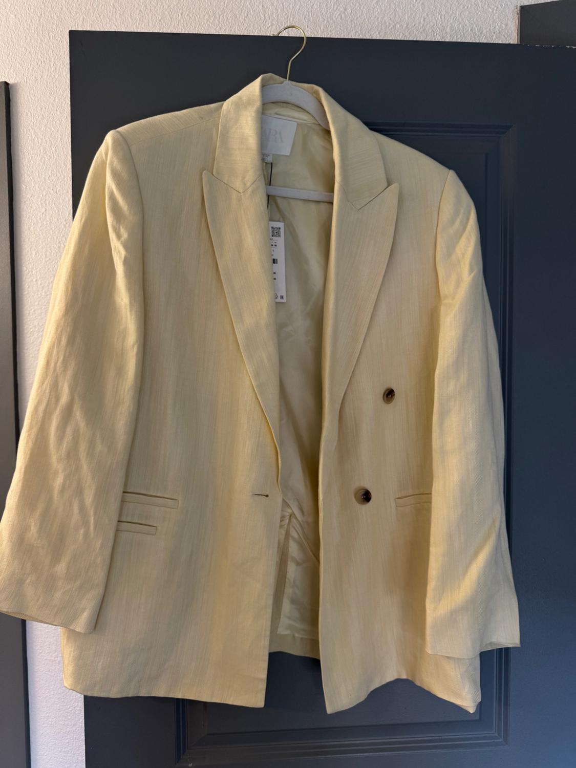 Yellow jacket hanging on a dark door, with a wide lapel and two brown buttons. Label visible inside.