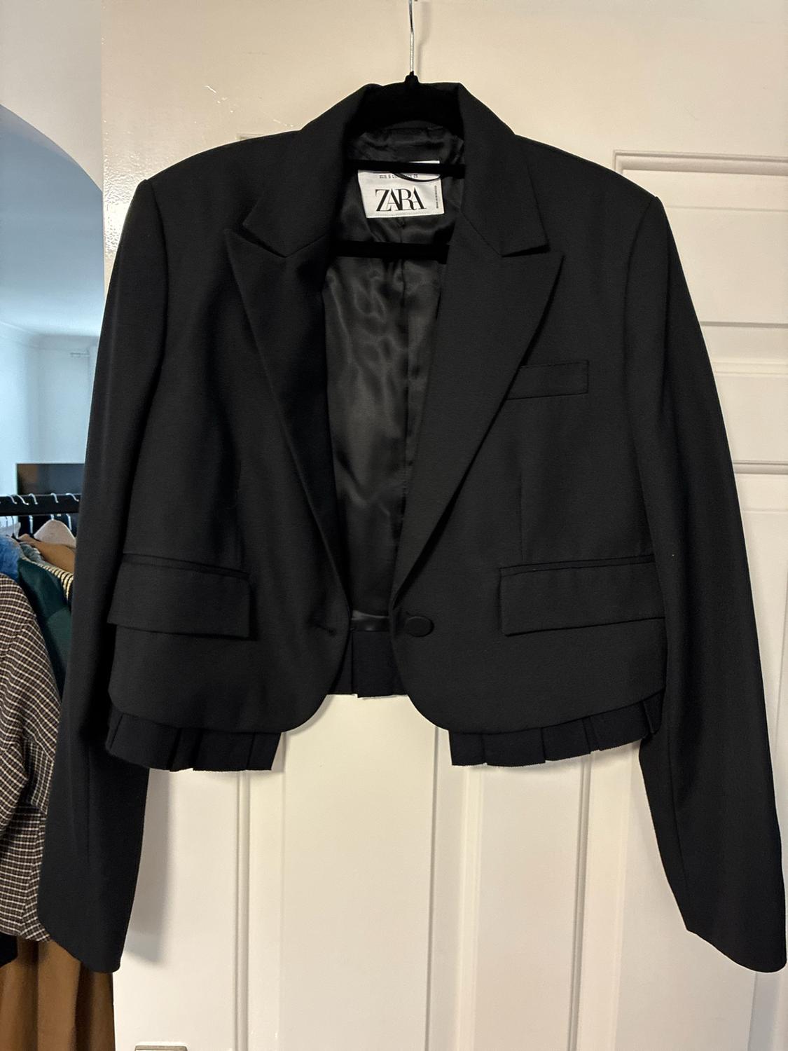 Short black jacket with pleated detailing at the hem and pockets, hanging on a white door. Internal label with the text ZARA.