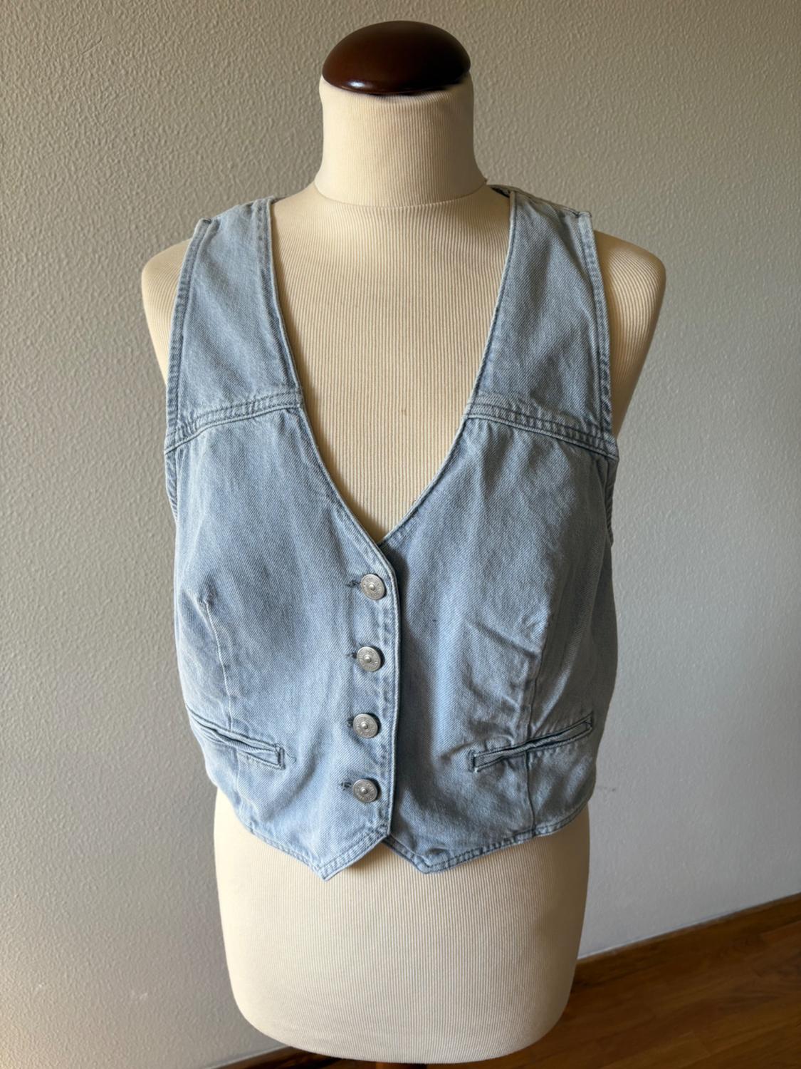 Light blue denim waistcoat with front buttons and a V-neck, displayed on a beige mannequin in front of a grey textured wall.