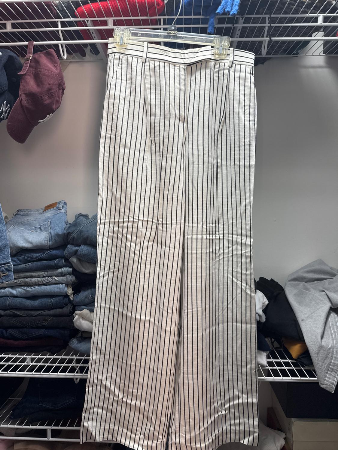 White trousers with vertical black stripes hanging on a transparent hanger, surrounded by folded clothes and caps on a shelf.