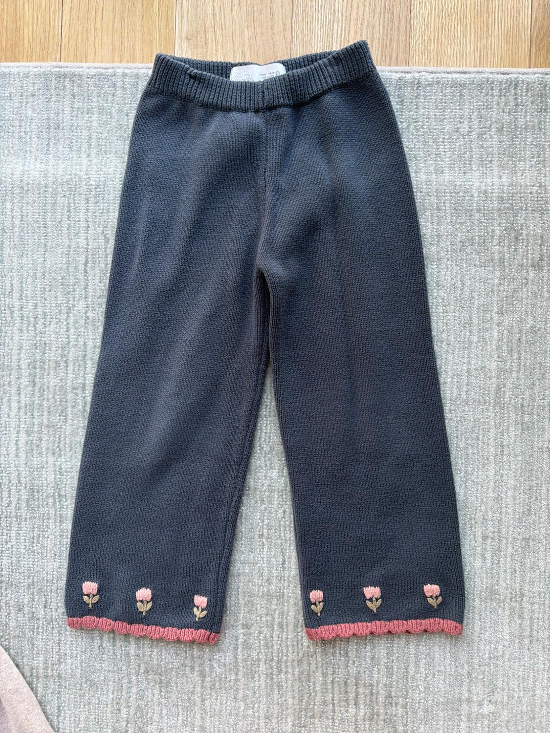 Black knitted trousers with pink floral embroidery on the hem, on a grey rug and light wood floor.