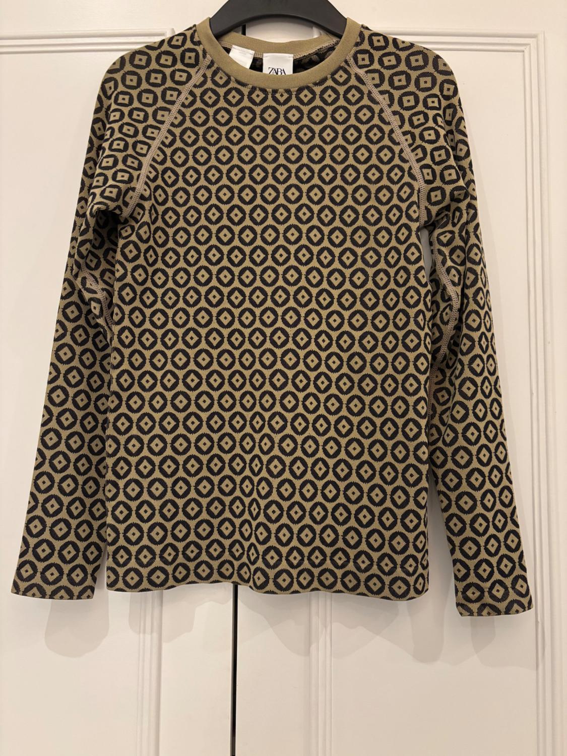 Long-sleeved jumper with a geometric pattern in beige and black tones, hanging on a hanger in front of a white door.