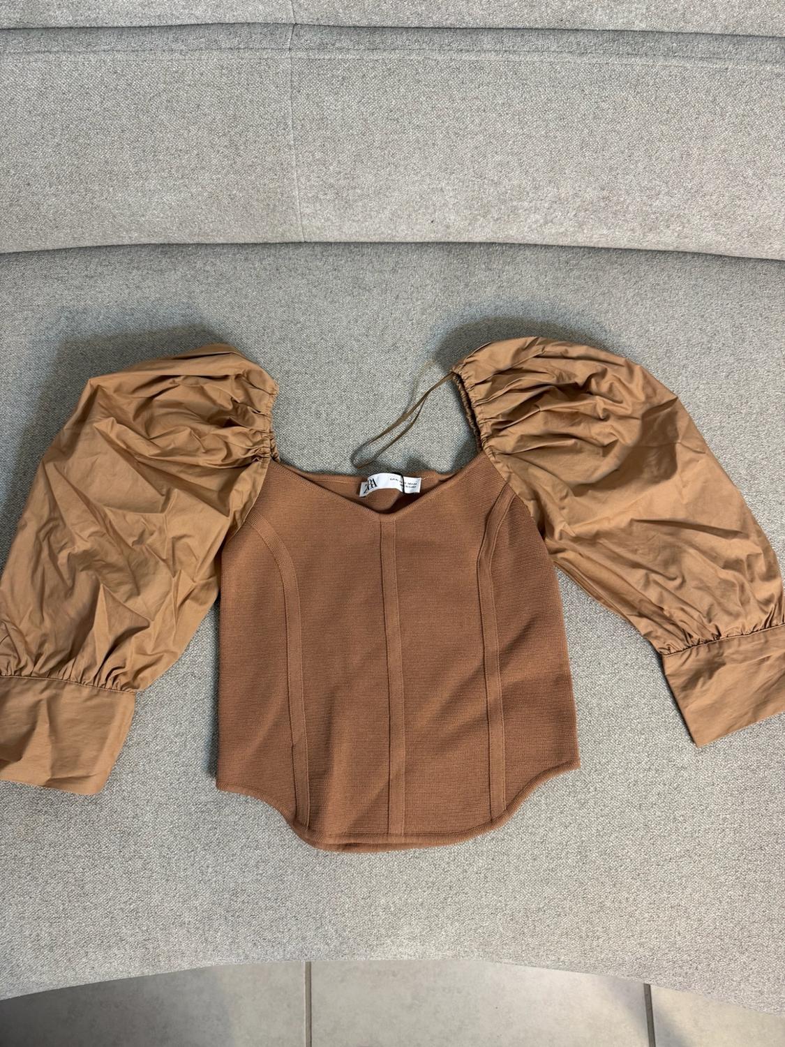 Brown top with a knitted body, vertical seams and puff sleeves on a grey textile surface. ZARA text on the label.