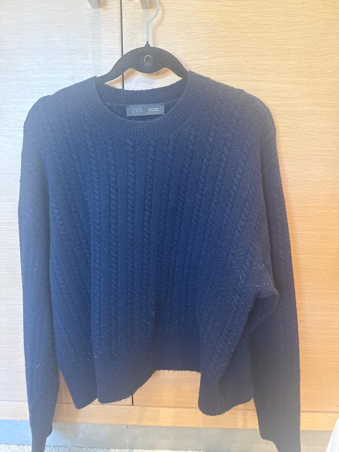 Navy cable-knit jumper hanging on a black hanger in front of a light wooden background. Visible label: Zara.