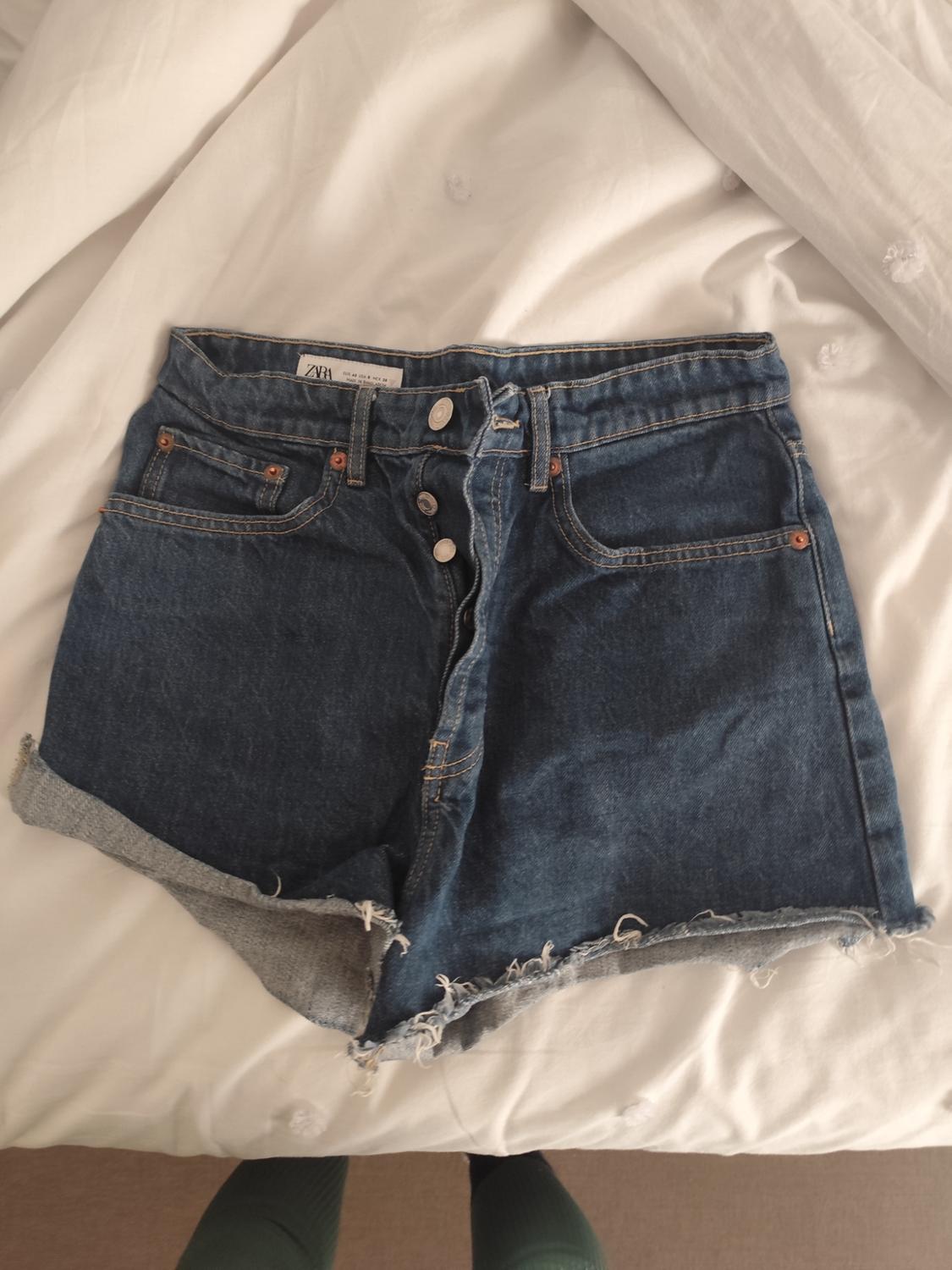 Blue denim shorts with visible buttons and frayed hem on a white bed. Visible label: ZARA.