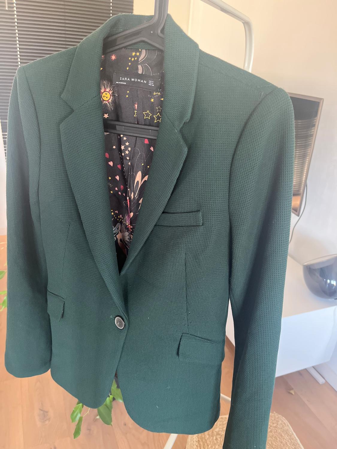 Dark green jacket hanging on a hanger, with a star and sun print lining. Visible label: "ZARA WOMAN". Modern room background.