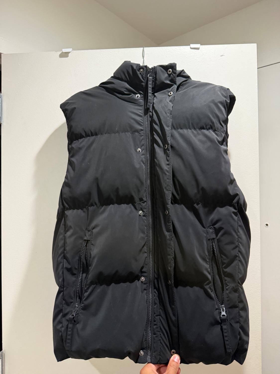 Here’s the rephrased sentence: A black puffer vest with a hood, a zipper front, and snap buttons; it’s hanging from a white-colored door and is held by a hand from the bottom.