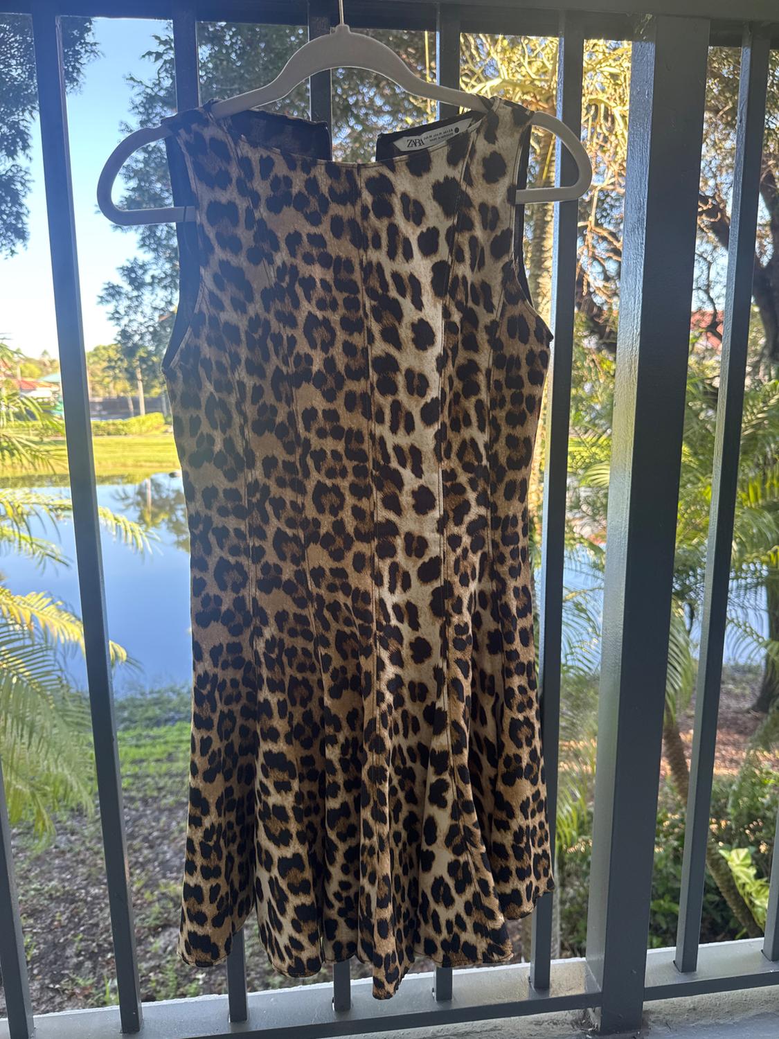 Animal print dress hanging on a hanger, with a natural landscape background and metal railing. Visible tag: Zara.