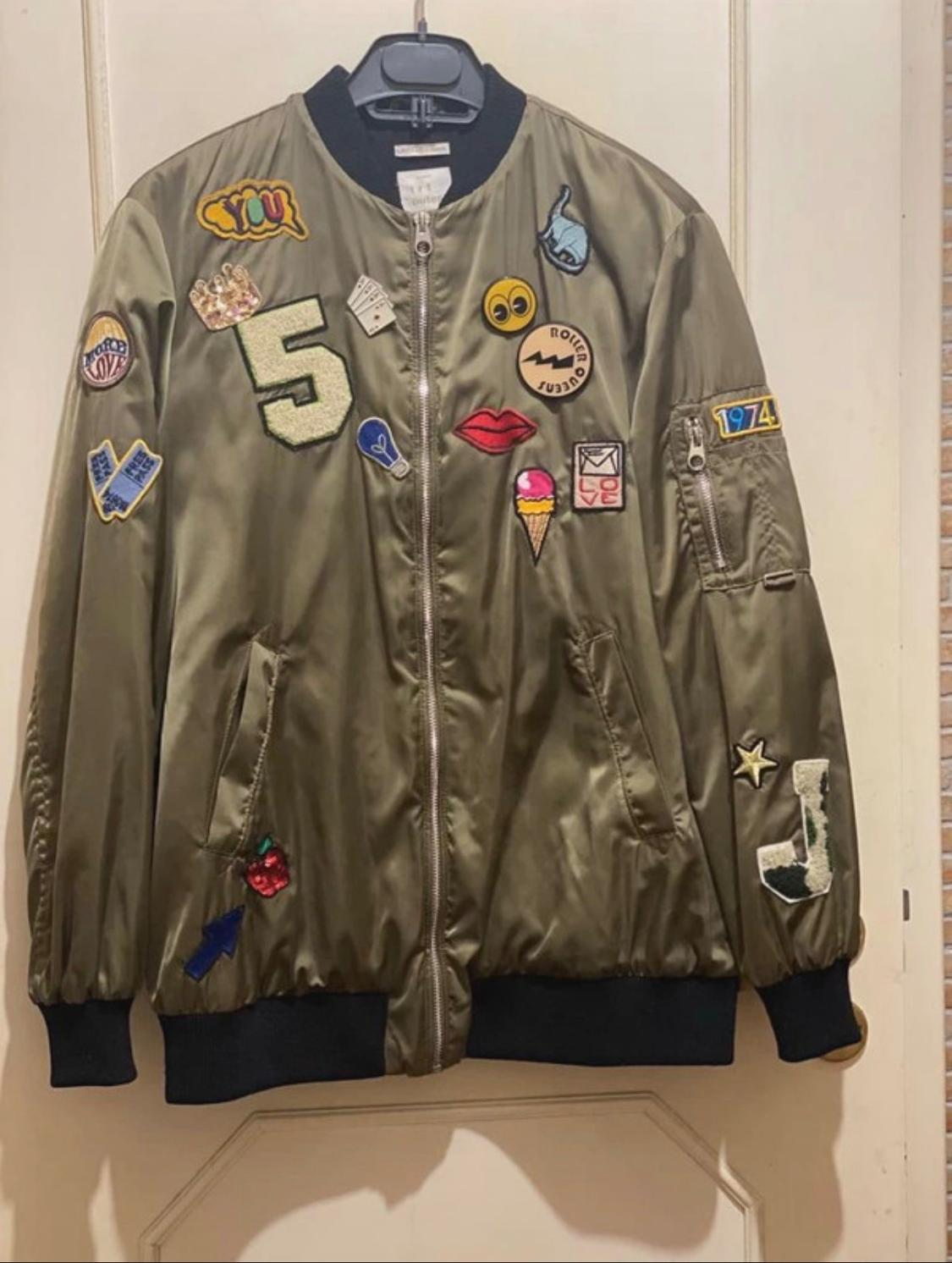Green bomber jacket embellished with colourful number, lip, ice cream, and "LOVE" text patches. Features black detailing on the cuffs and collar.