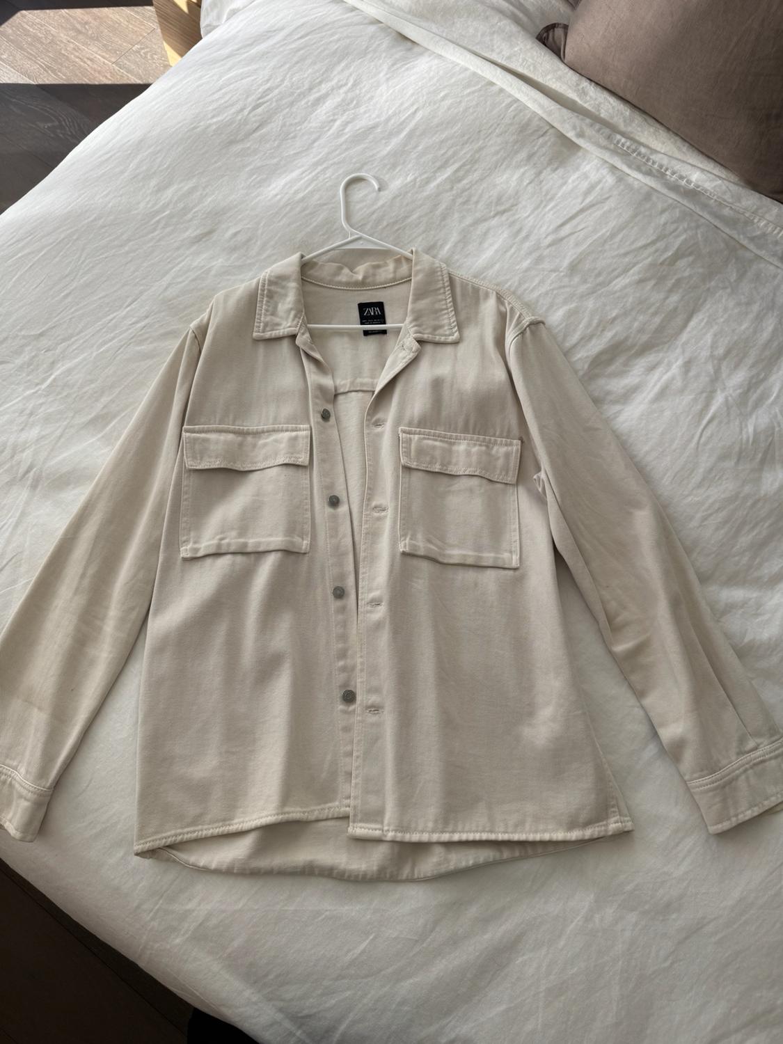 Beige button-up shirt with two chest pockets and collar, hanging on a white hanger over a white sheet. Visible Zara label.