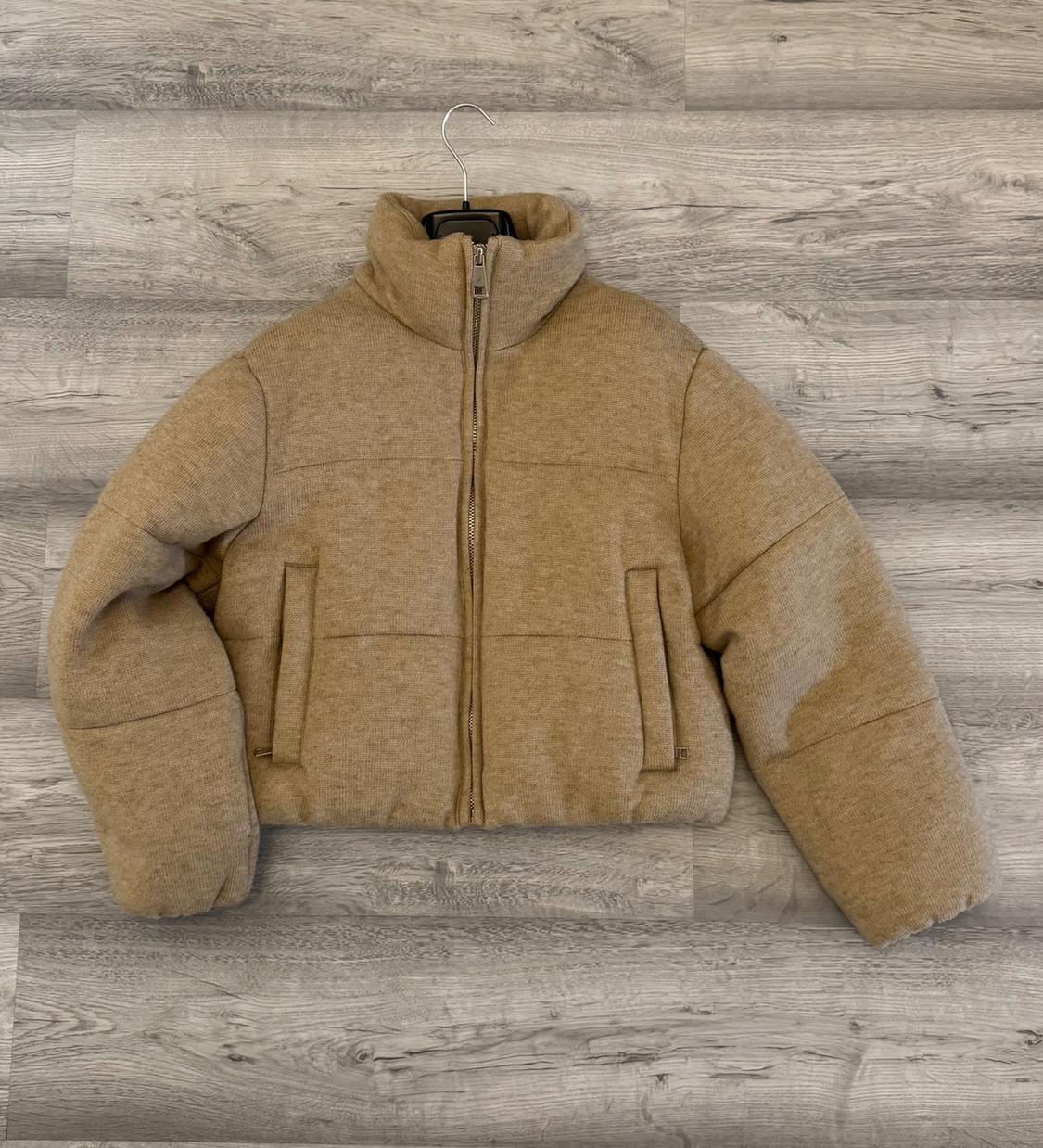 Beige quilted jacket hanging on a hanger against a grey wooden background. Zip-up design with a high neck.