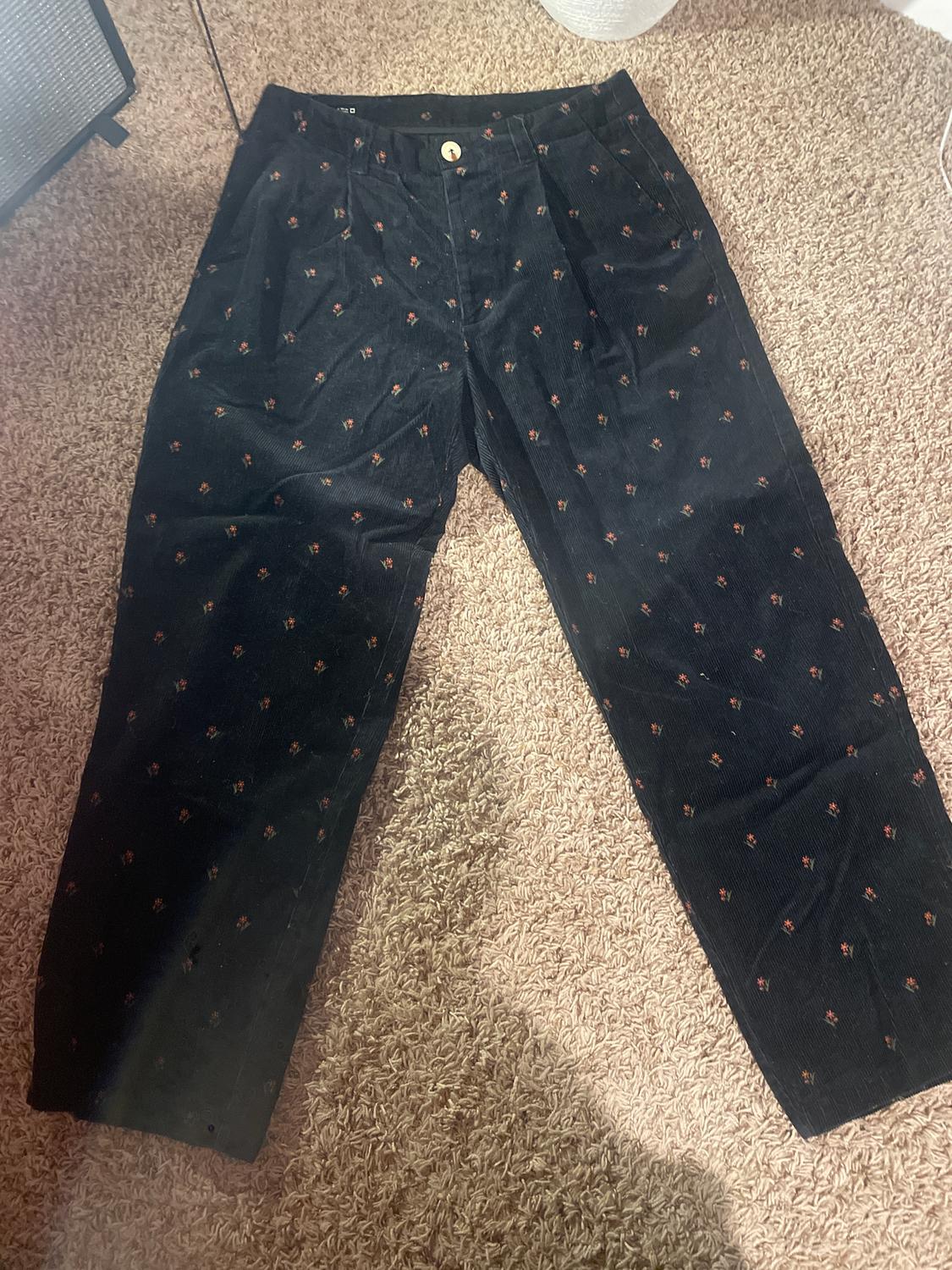 Black corduroy trousers with a small red and yellow floral print, on a beige rug. Gold button visible at the waist.